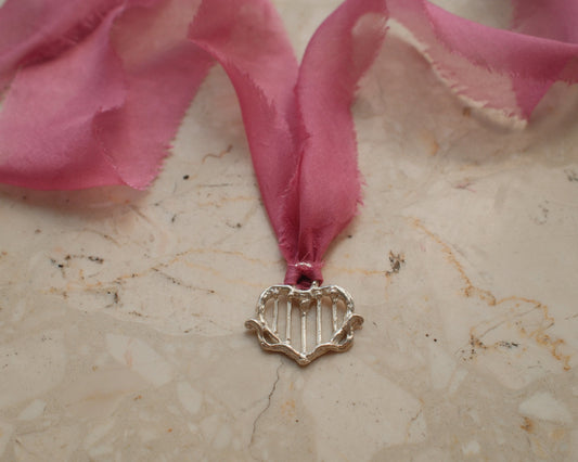 A silver-toned metal heart-shaped pendant with vertical bar details, hanging from a sheer pink ribbon on a beige marble surface.