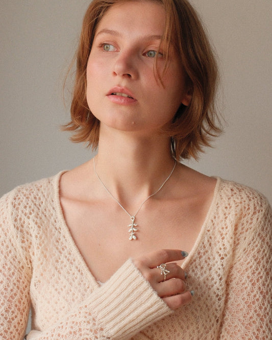 A woman with short light brown hair wearing a cream-colored open-knit sweater and a delicate silver necklace with a small tree pendant, posing against a neutral beige background.