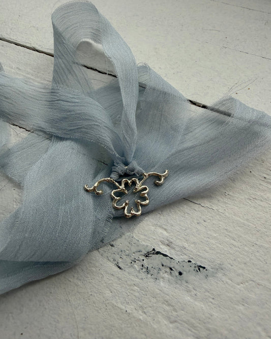 A light blue sheer silk ribbon gathered with a small silver flower charm, laid on a distressed white wooden surface.