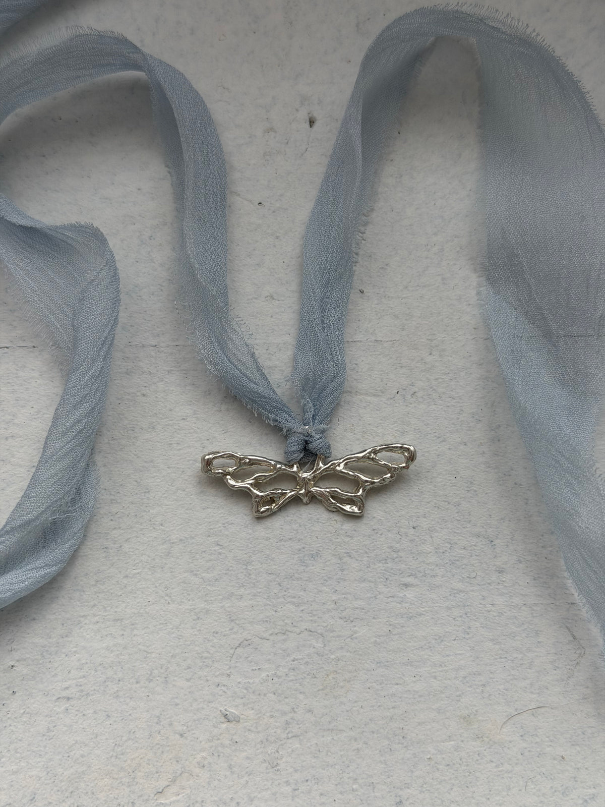 A silver butterfly-shaped pendant or charm threaded onto a sheer, light blue ribbon, laid flat on a textured off-white surface.